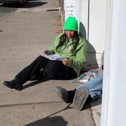a person sitting on the sidewalk leaning against the white building writing on a clipboard